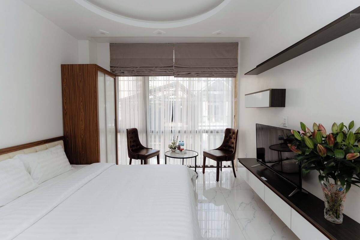 A hotel room with a bed, a TV, a small table with chairs, and a balcony with curtains.