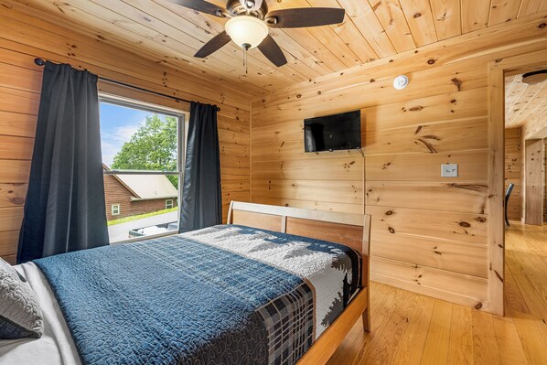 Habitación de cabaña de madera con una cama, ventilador de techo, ventana con cortinas y televisor montado.