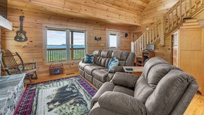 Interior acogedor de una cabaña con un sofá grande de color gris, techo de madera y vistas a través de las ventanas.