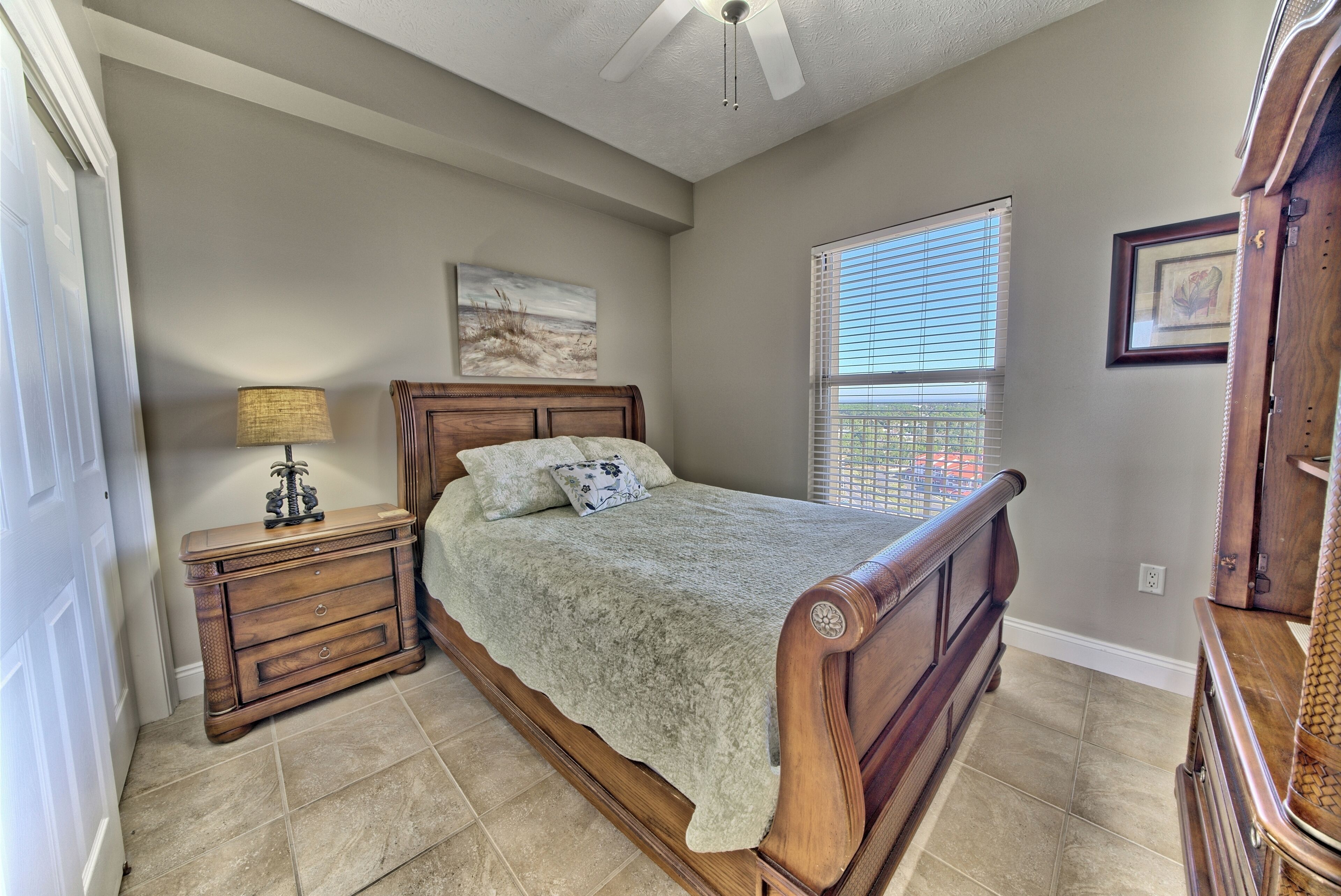 A bedroom with a wooden bed, a nightstand, a lamp, and a window with blinds.