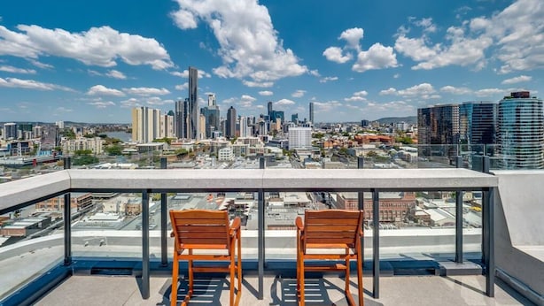 Una terraza en la azotea con dos sillas de madera y una mesa, con vistas a un horizonte urbano de edificios modernos y un cielo despejado.