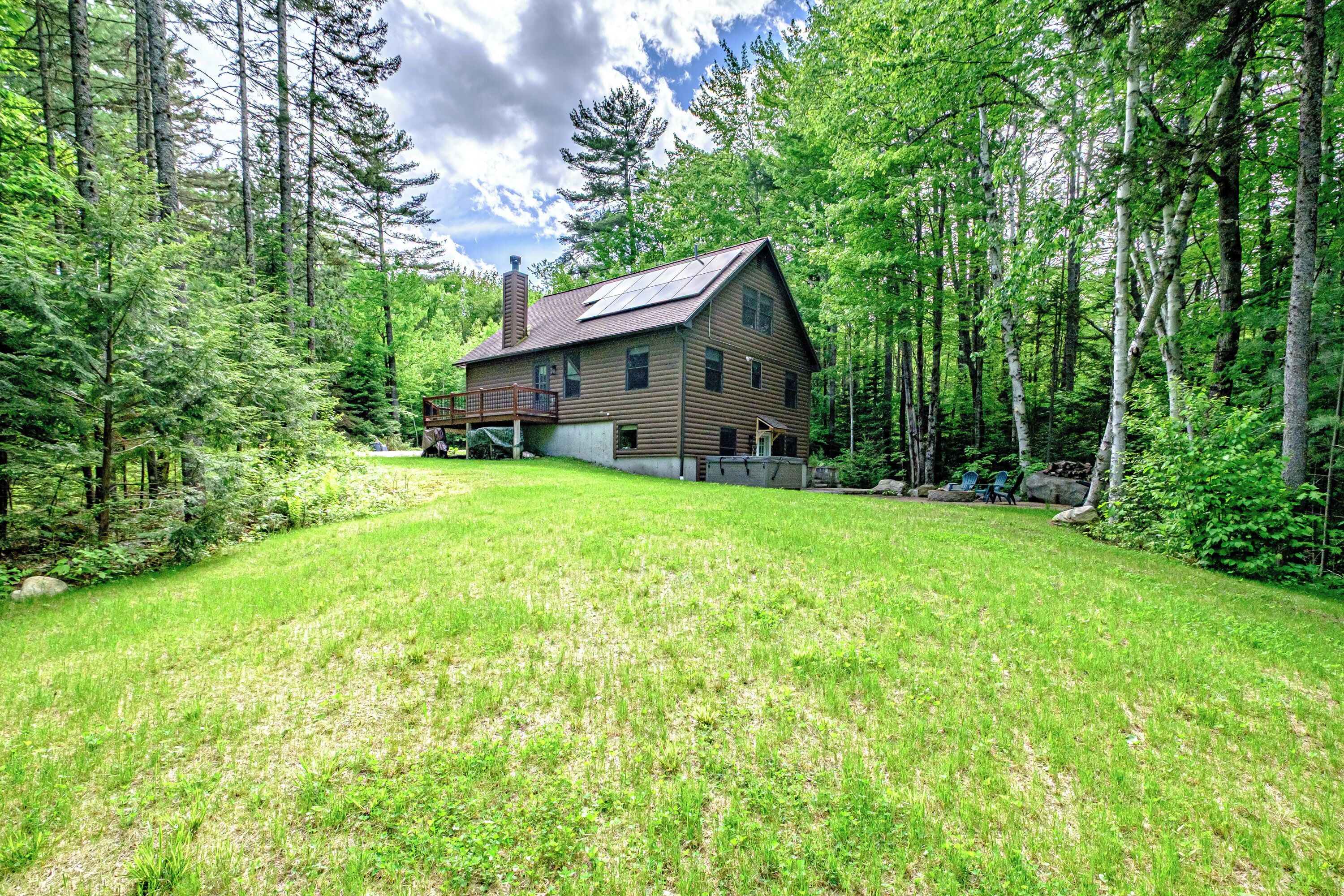 A two-story log cabin with a deck and solar panels, surrounded by dense forest.