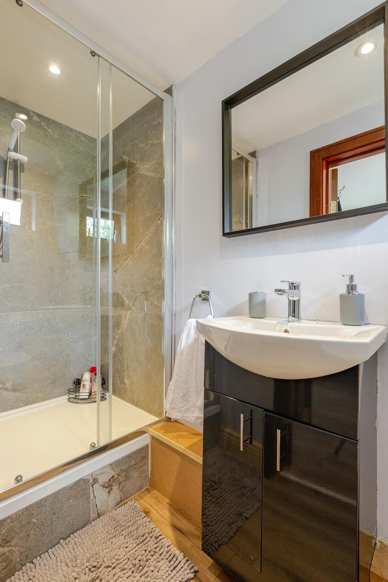 Un baño moderno con ducha envuelta en vidrio, un lavabo blanco con dos grifos y un espejo grande sobre el lavabo.