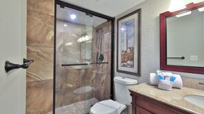 A bathroom with a glass-enclosed shower, a toilet, a sink with a granite countertop, and a framed picture on the wall.