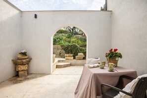 Un espace patio aménagé avec une table et des chaises, un banc en pierre et une vue sur un jardin, accessible par un arc.