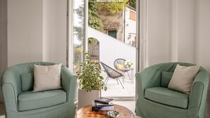 Een kamer met twee groene fauteuils, een ronde salontafel met boeken en een schaal, en uitzicht op een gebouw door een raam.