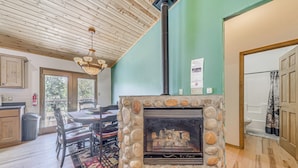 Une cheminée en pierre avec une hotte, un coin repas avec une table et des chaises, et une vue sur l’extérieur par une porte.