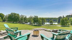 Eine Terrasse mit Adirondack-Stühlen und einem Feuerkorb mit Blick auf einen See und einen Wald.