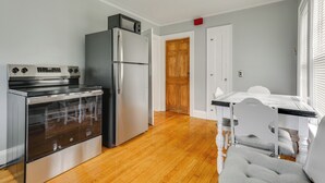Una cocina con horno empotrado y refrigerador, pisos de madera, una mesa de comedor con sillas, y una puerta de madera al fondo.