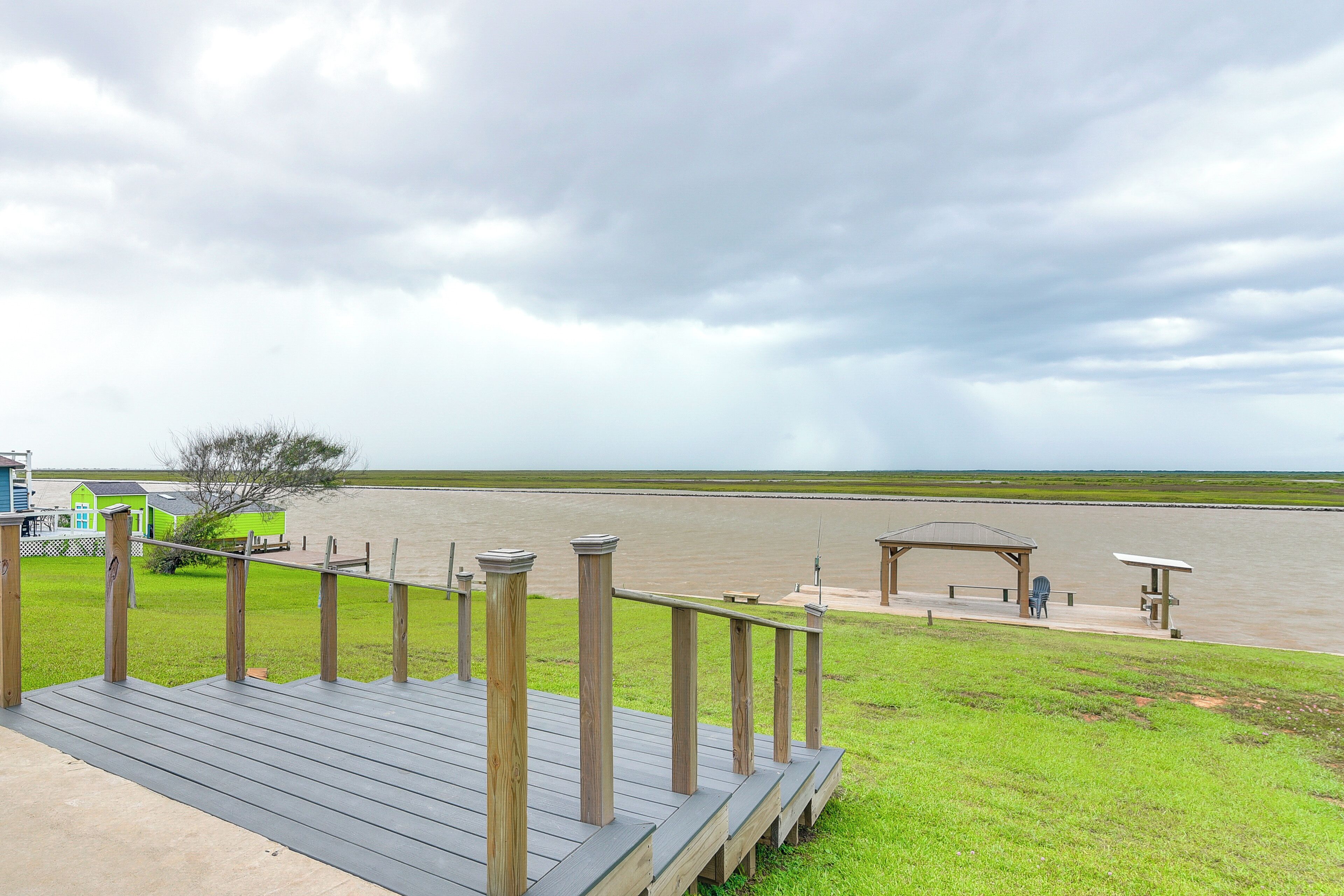 A wooden deck with a railing overlooking a body of water, a grassy area, and a small structure with a roof.