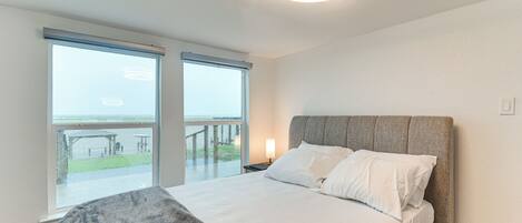 A bedroom with a bed, a nightstand, a lamp, and a view of the outdoors through large windows.