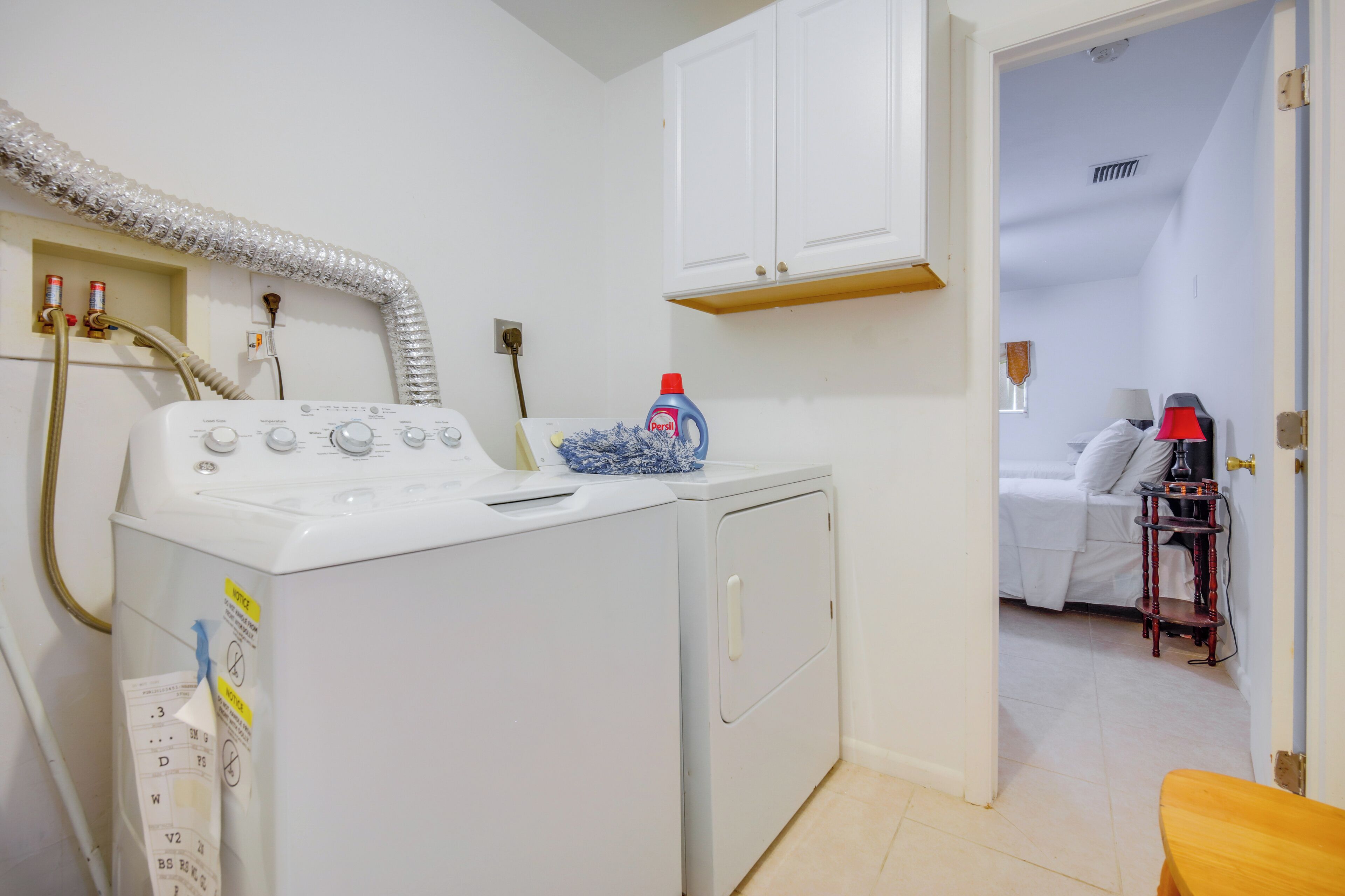 Il y a un espace buanderie avec un lave-linge, un sèche-linge et une étagère où sont rangés les produits de lessive. La pièce adjacente contient un lit et une table de chevet.