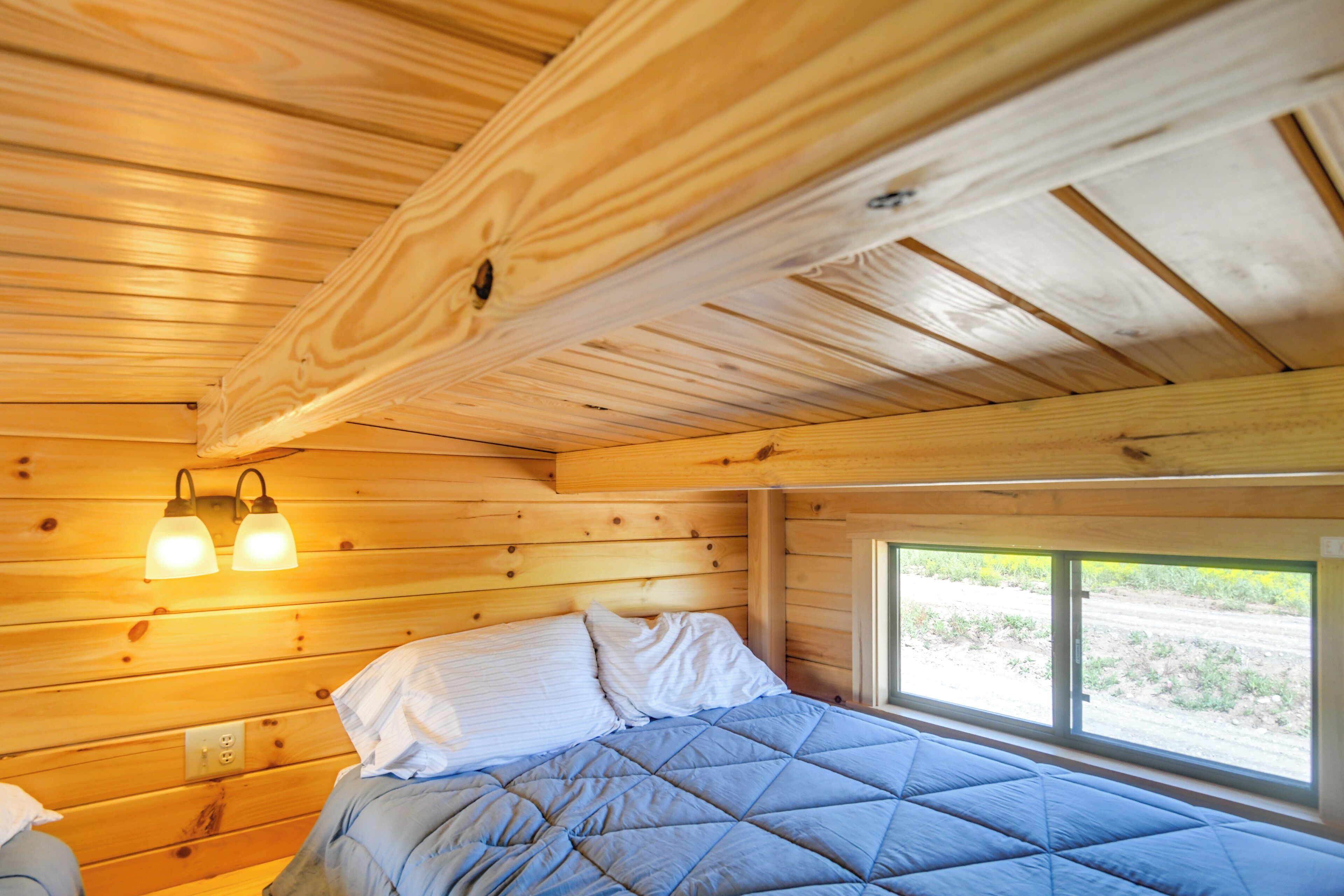 A wooden cabin interior with a bed, wooden walls, and a window with a view of the outdoors.
