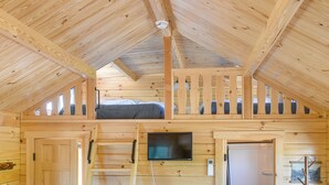 A wooden cabin interior with a loft bed, a television mounted on the wall, and a door leading to another room. - Teton Mtn Views: Peaceful Riverfront Cabin! (Ashton)