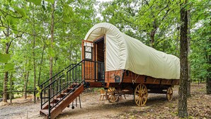 A covered wagon with a wooden staircase leading up to an open door, surrounded by trees in a forest setting. - Covered Wagon Vacation Rental in Broken Bow! (Broken Bow)