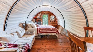 A cozy, circular tent-style room with a wooden floor, a bed with a red and white quilt, a sofa, and a wooden chair. - Covered Wagon Vacation Rental in Broken Bow! (Broken Bow)