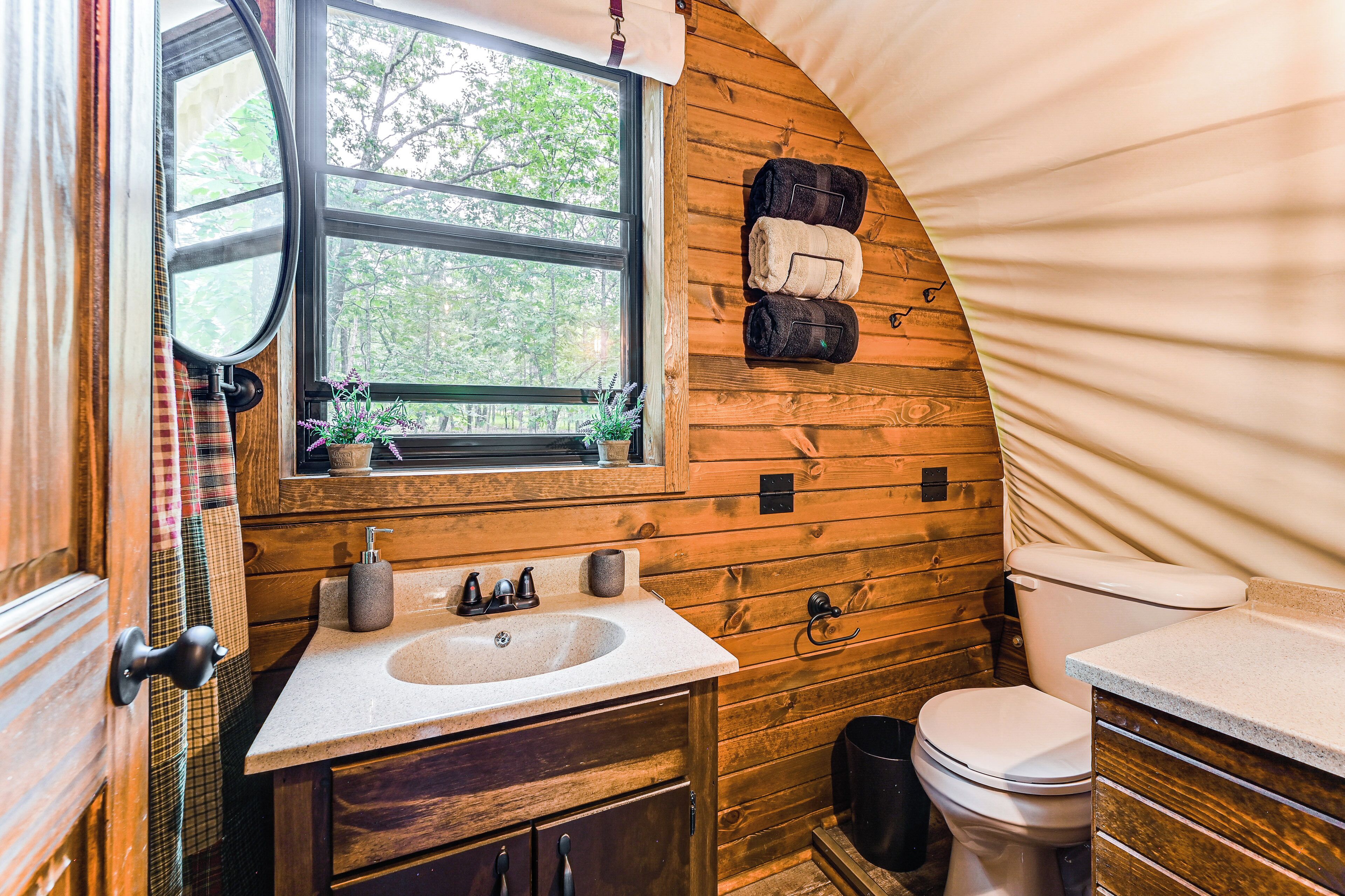 Ein kleines, rustikales Badezimmer mit Holzvertäfelung, einem Fenster mit Blick auf Grünflächen, einem Waschbecken mit Wasserhahn, einer Toilette und einem Handtuchhalter mit Handtüchern.