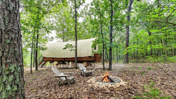 A rustic campsite with a covered wagon, wooden chairs, and a fire pit surrounded by trees. - Covered Wagon Vacation Rental in Broken Bow! (Broken Bow)
