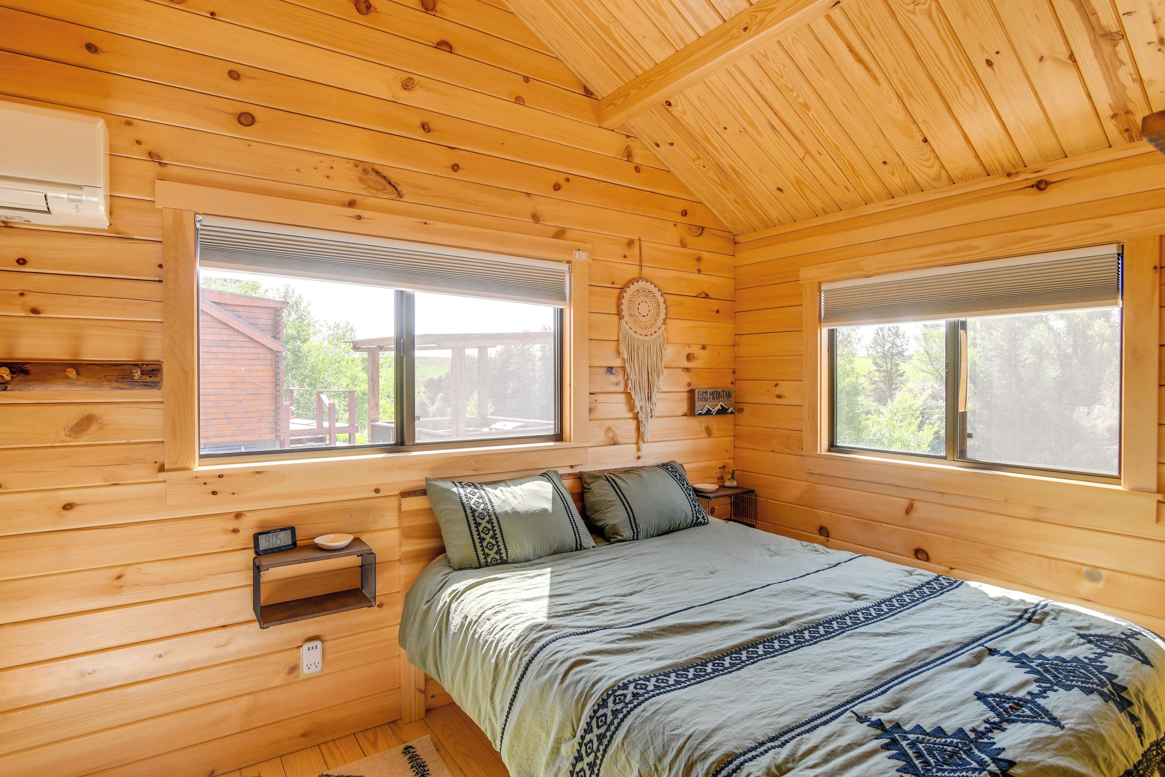 Un dormitorio de cabaña de madera con dos ventanas, una cama con ropa de cama estampada, una mesita de noche y un atrapasueños en la pared.