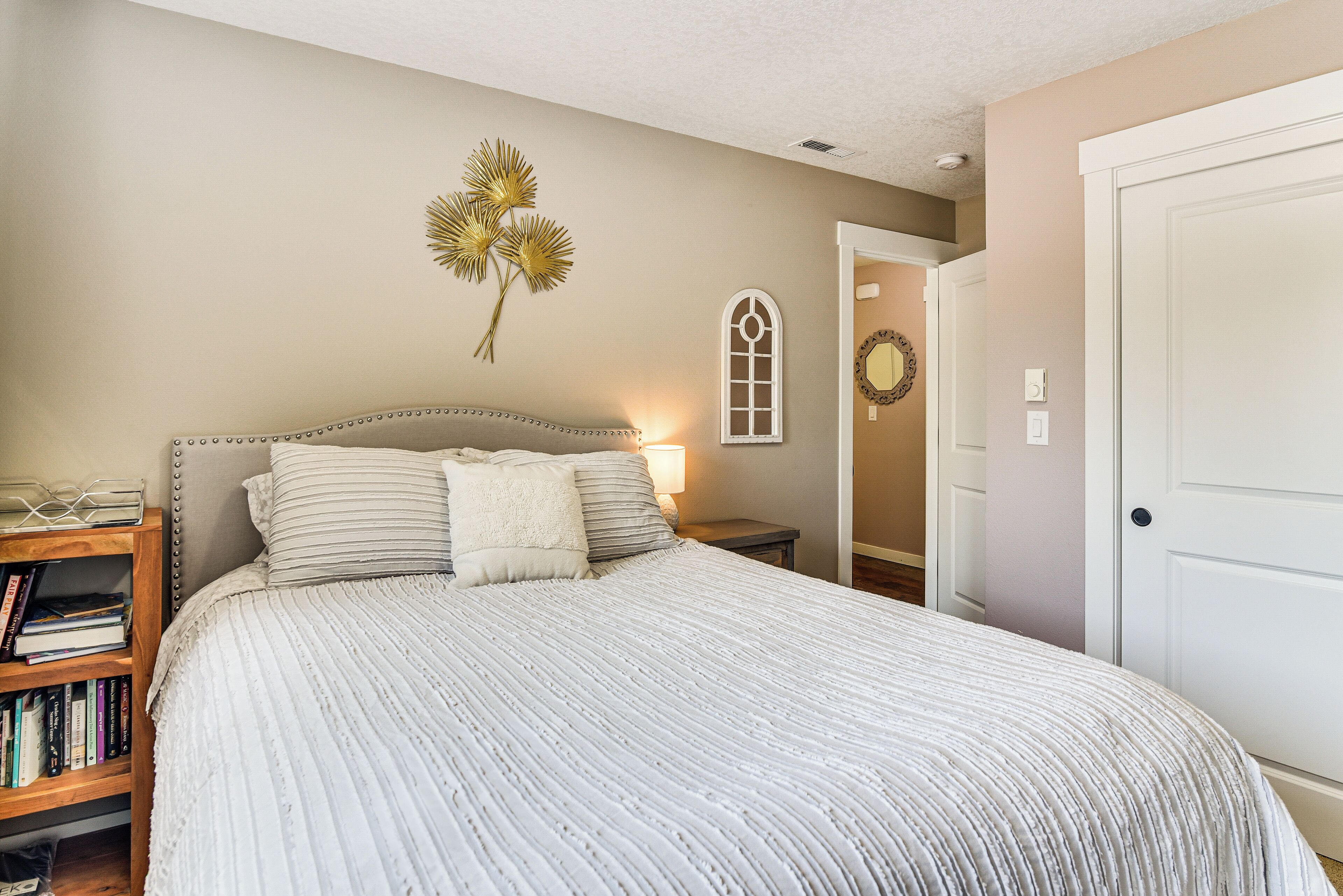 A bedroom with a large bed, a bedside table with a lamp, a bookshelf, and a door to another room.