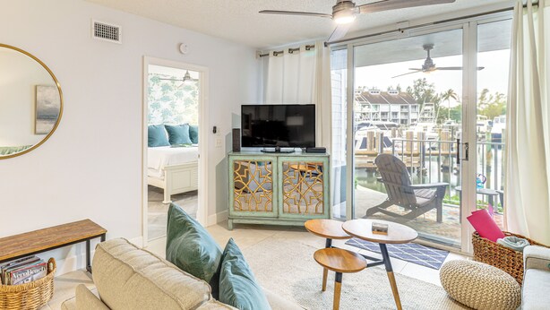 Ein Wohnzimmer mit einer Couch, einem Fernseher auf einem Schrank, einem Deckenventilator und Blick auf einen Balkon mit einem Stuhl.
