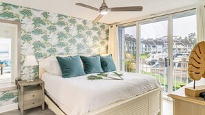 Ein Schlafzimmer mit Bett, Nachttisch, Lampe und Blick auf Boote durch große Fenster.