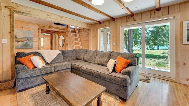 A cozy living room with a sectional sofa, a wooden coffee table, and a large window offering a view of the outdoors. - Dock + Kayaks: Annabessacook Lake Retreat! (Winthrop)