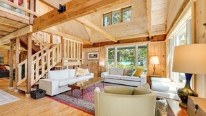 A spacious living room with wooden beams, a staircase, and large windows offering a view of the outdoors. - Dock + Kayaks: Annabessacook Lake Retreat! (Winthrop)