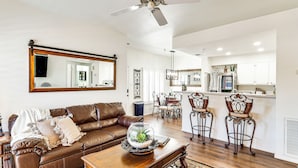 A spacious living room with a brown leather sofa, a coffee table with a glass dome containing a plant, and a dining area with metal chairs and a wooden table. - Walk to Rotary Park: LHC Condo w/ Private Patio (Lake Havasu City)