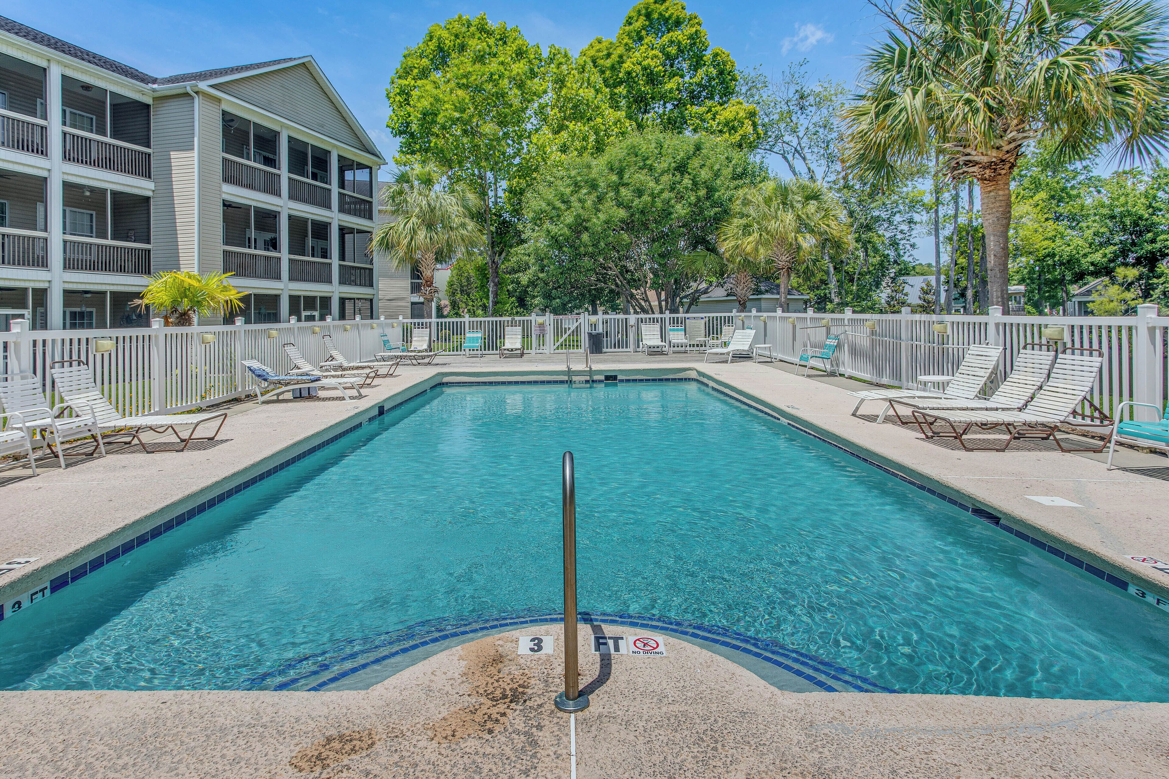 A rectangular pool with a white fence, lounge chairs, and a building with balconies in the background.