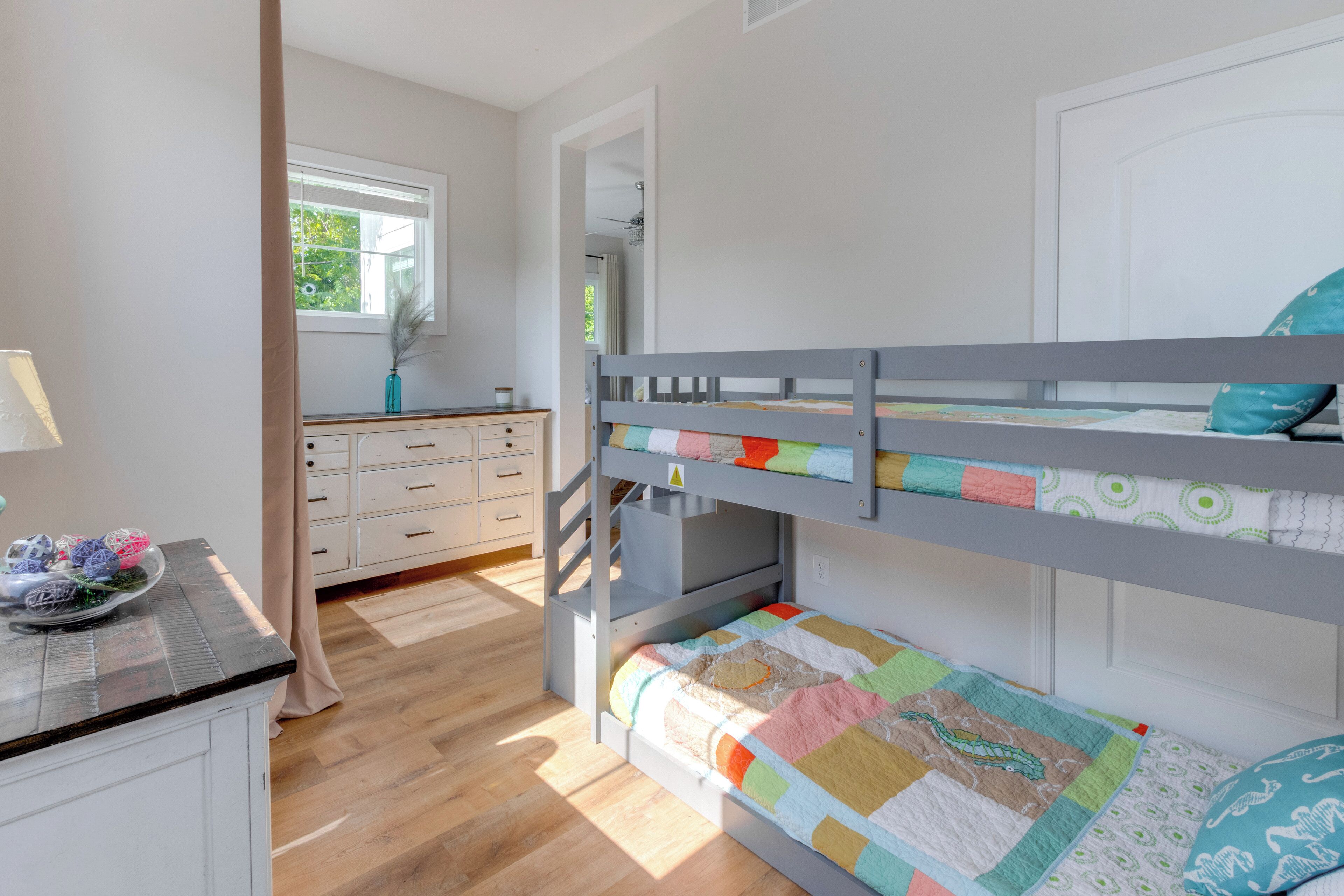 A bunk bed room with a wooden floor, a desk with a lamp, and a window with a view of greenery.