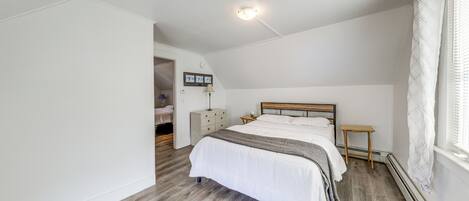 A bedroom with a wooden floor, a bed with white bedding, a bedside table, a nightstand, a lamp, a heater, and a window with curtains.