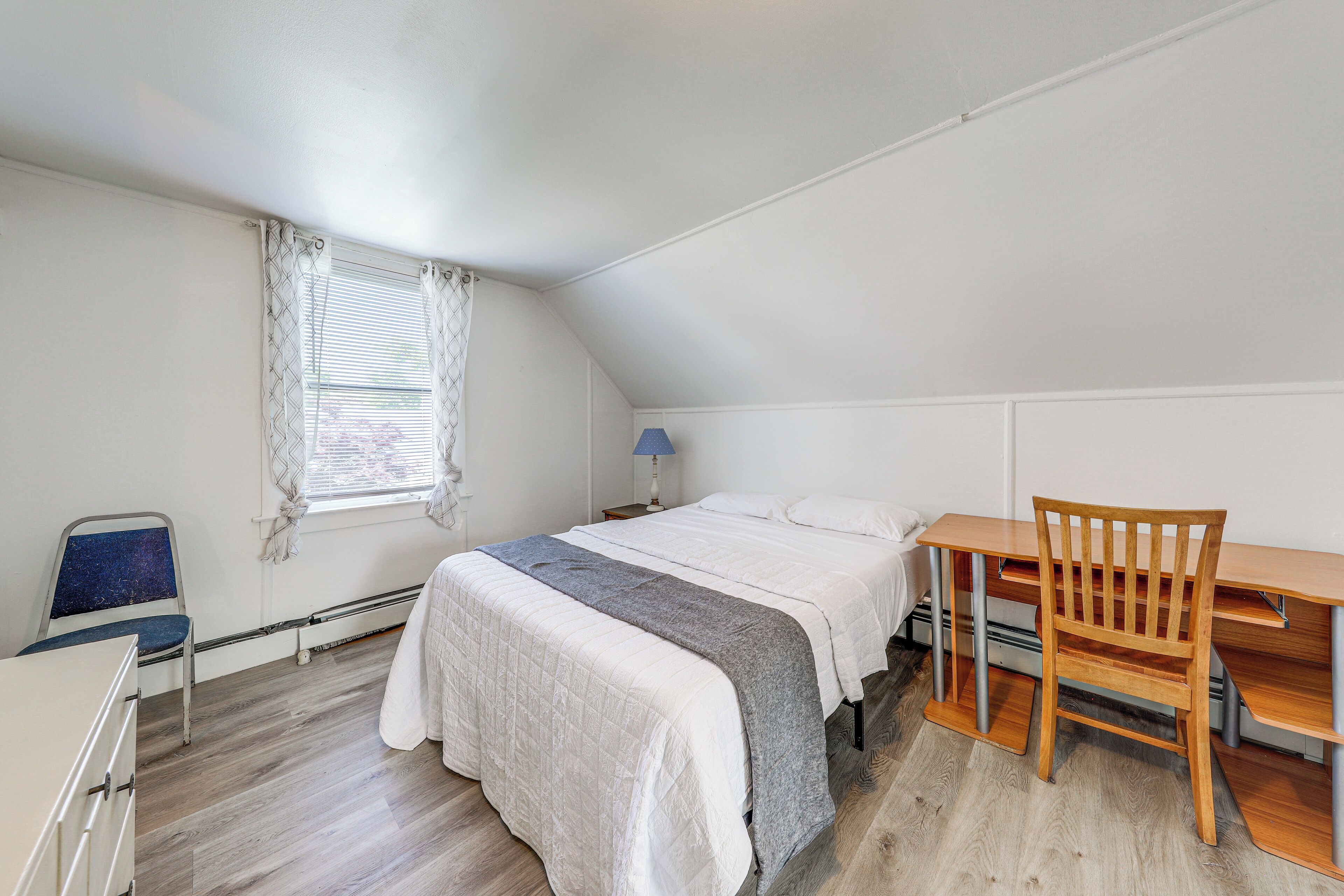 A bedroom with a bed, a desk, a chair, a window with curtains, and a radiator.