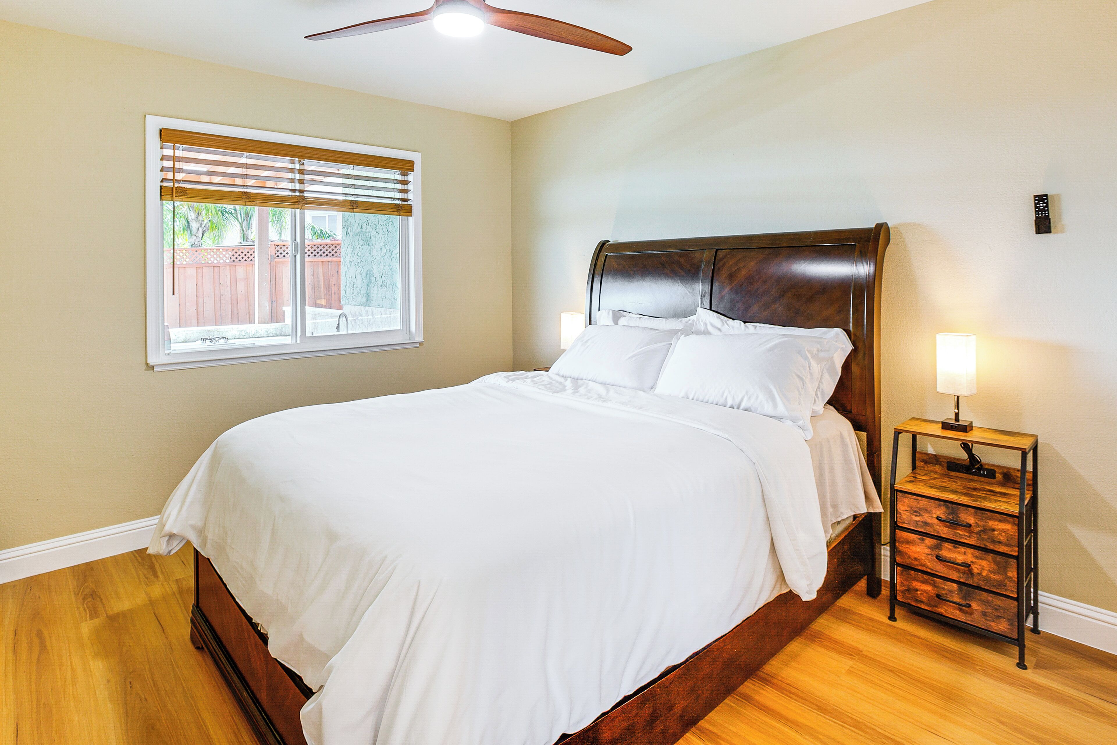 Une chambre à coucher avec un lit en bois, une table de chevet avec des tiroirs, un ventilateur de plafond et une fenêtre avec des stores.
