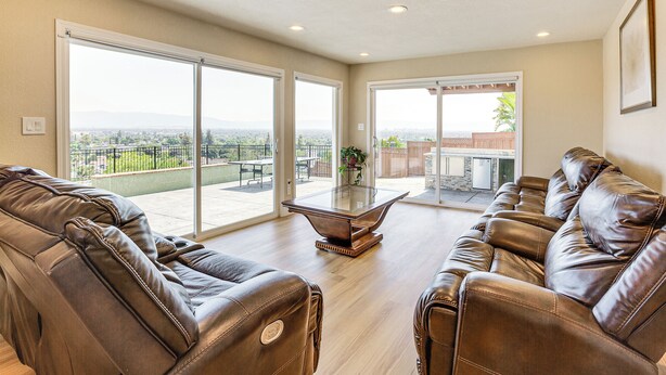 A spacious living room with leather sofas, a glass coffee table, and large sliding glass doors leading to a balcony with a city view.
