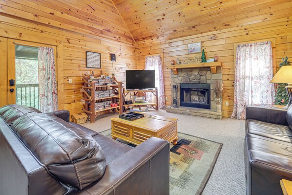 A cozy log cabin interior with a leather sofa, a television, a fireplace, and a wooden bookshelf. - Smokies Cabin on Douglas Lake w/ Resort Pool (Sevierville)