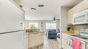 A modern kitchen with wooden cabinets, a white refrigerator, a microwave, and a stove. There is a dining area with a blue sofa and a ceiling fan. - 6 Mi to Lake Sumter Landing: Home w/ Perks! (The Villages)