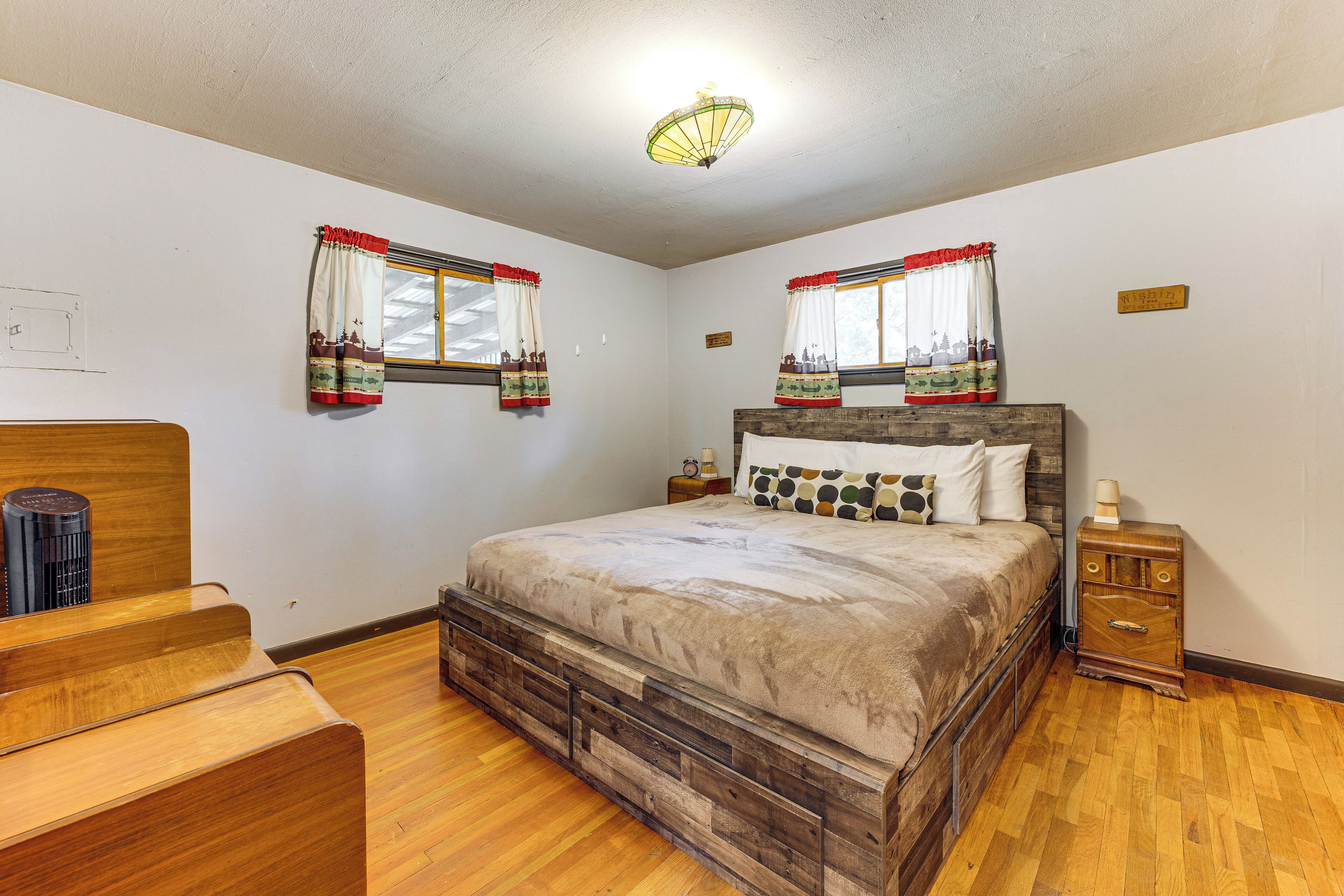 Un dormitorio con una cama de madera, dos mesitas de noche, un banco de madera y dos ventanas con cortinas.