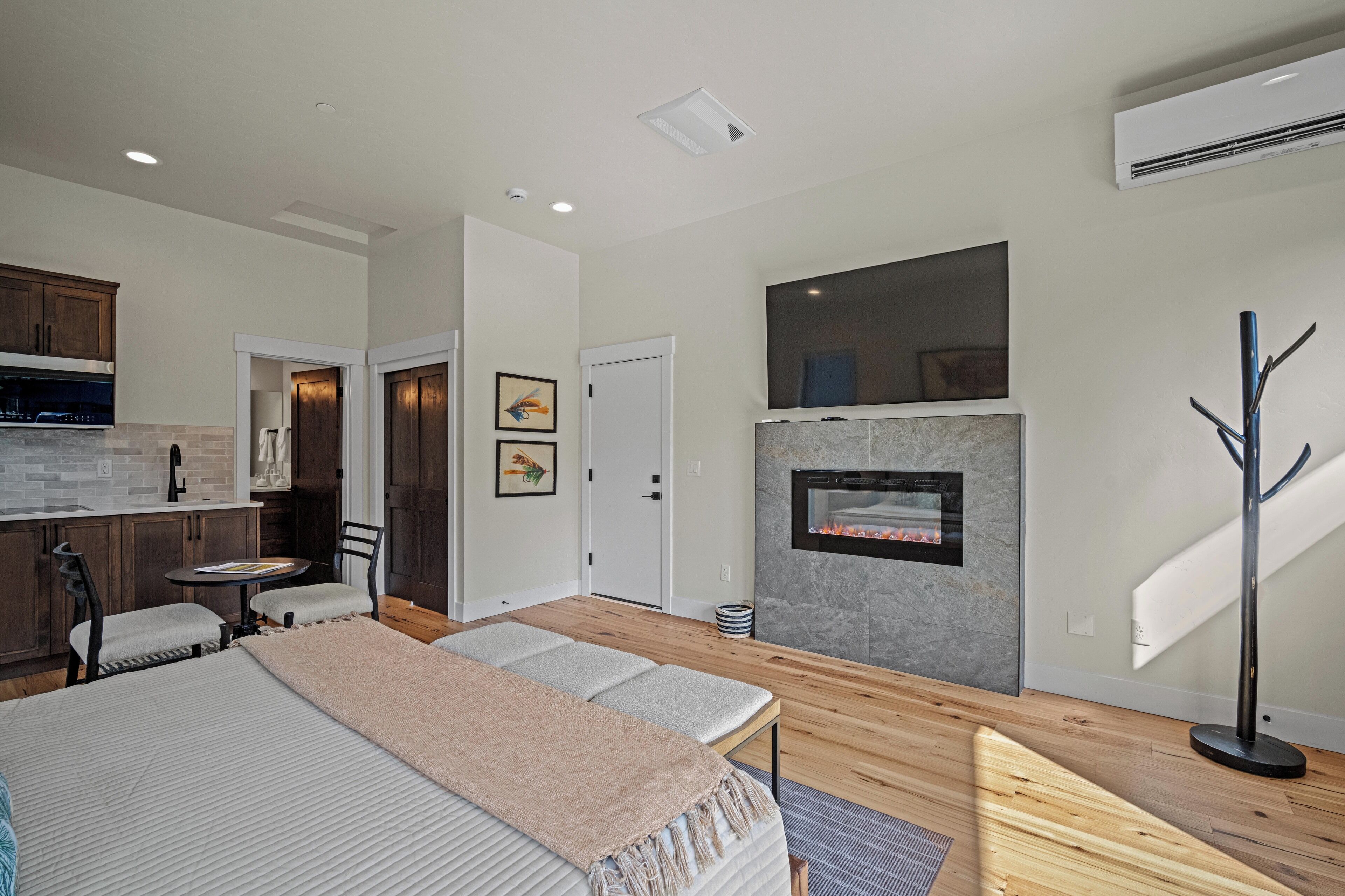 A modern hotel room with a bed, a small dining area, a fireplace, and a kitchen area.