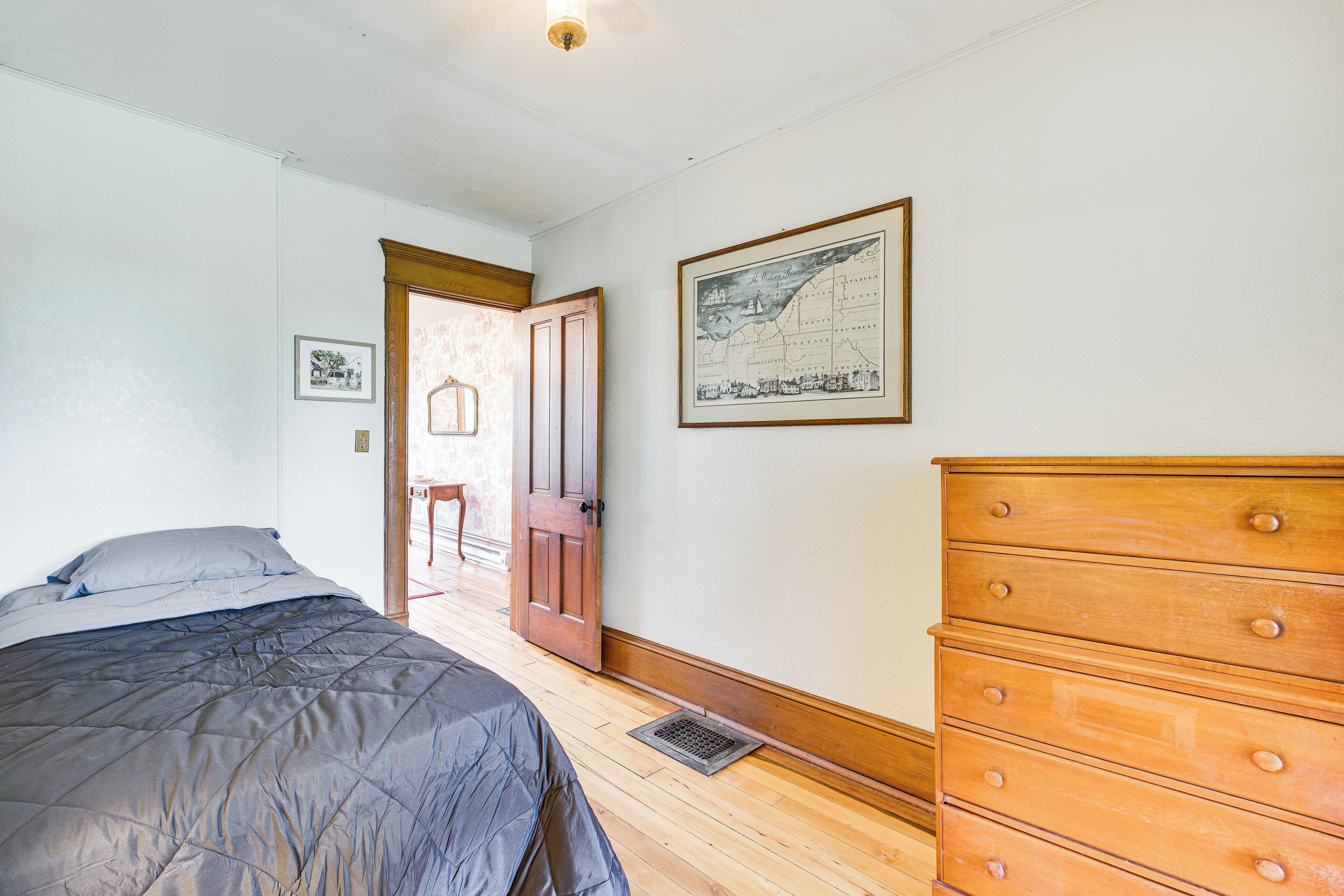 A bedroom with a bed, a wooden dresser, a framed map on the wall, and a door leading to another room.