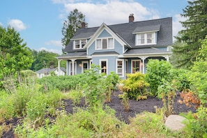 A large two-story house with a gabled roof, surrounded by lush greenery and a vegetable garden.