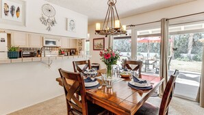A dining area with a wooden table set for four, surrounded by wooden chairs. A chandelier hangs above the table, and a vase with flowers is on the table.
