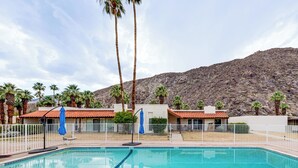 A swimming pool with a view of a mountainous landscape, surrounded by palm trees and a fenced area with a building in the background.