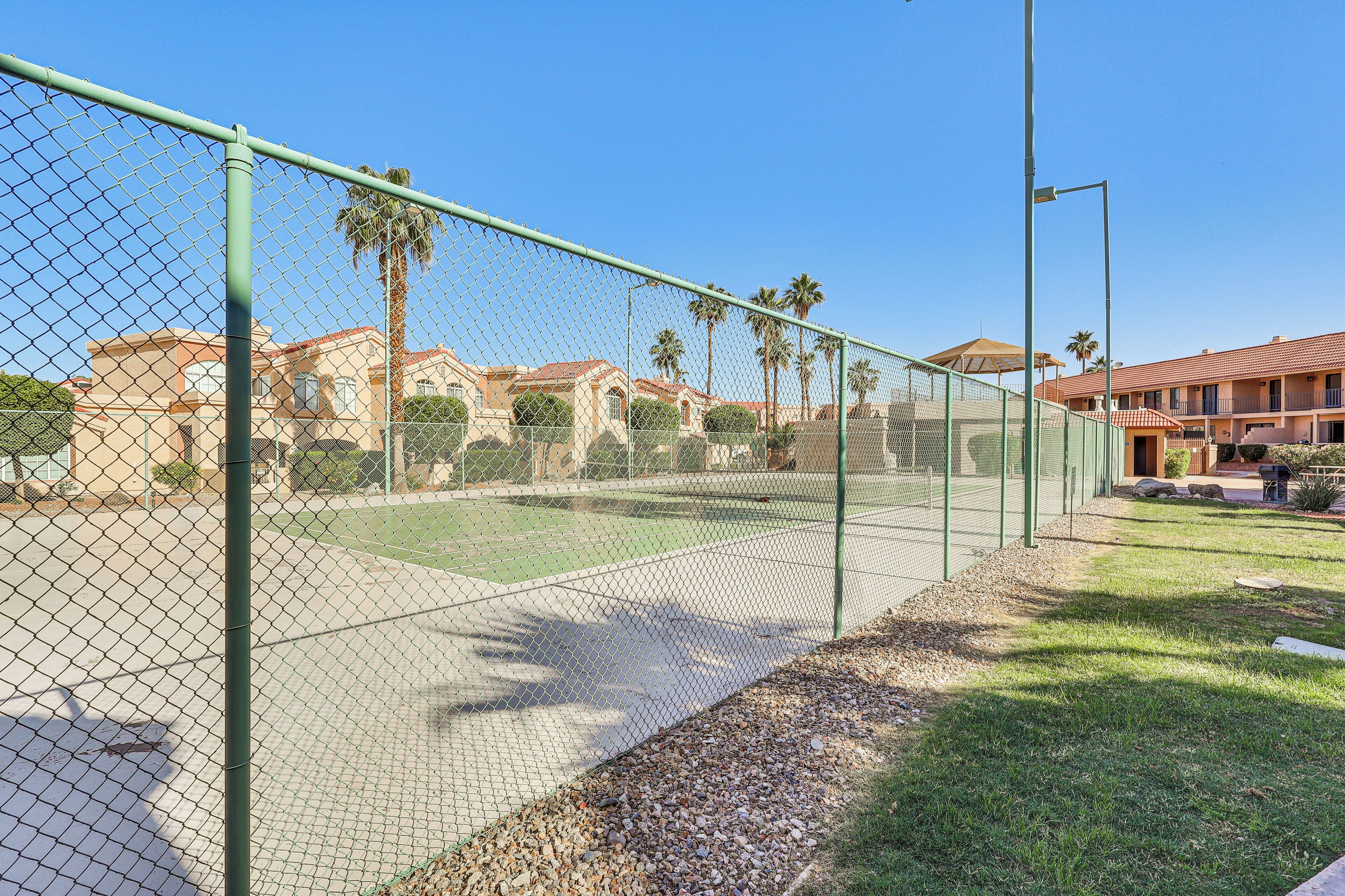 Una cancha de tenis cercada, con vista despejada a un edificio de techo rojo y palmeras al fondo.