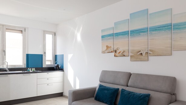 A modern kitchen with white cabinets and a grey countertop, a grey sofa with blue cushions, and a beach-themed wall art. - Casa Brezza Marina by Wonderful Italy (Olbia)