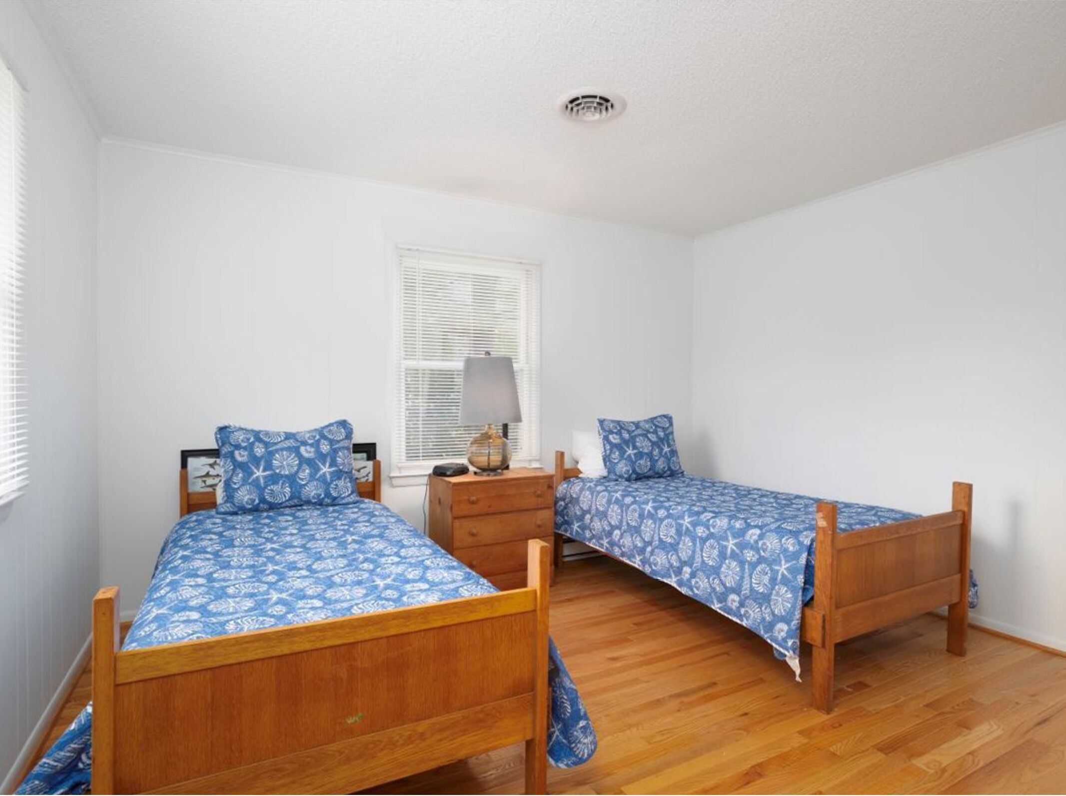 A bedroom with two wooden beds, white walls, a window with blinds, a bedside table with a lamp, and a wooden floor.