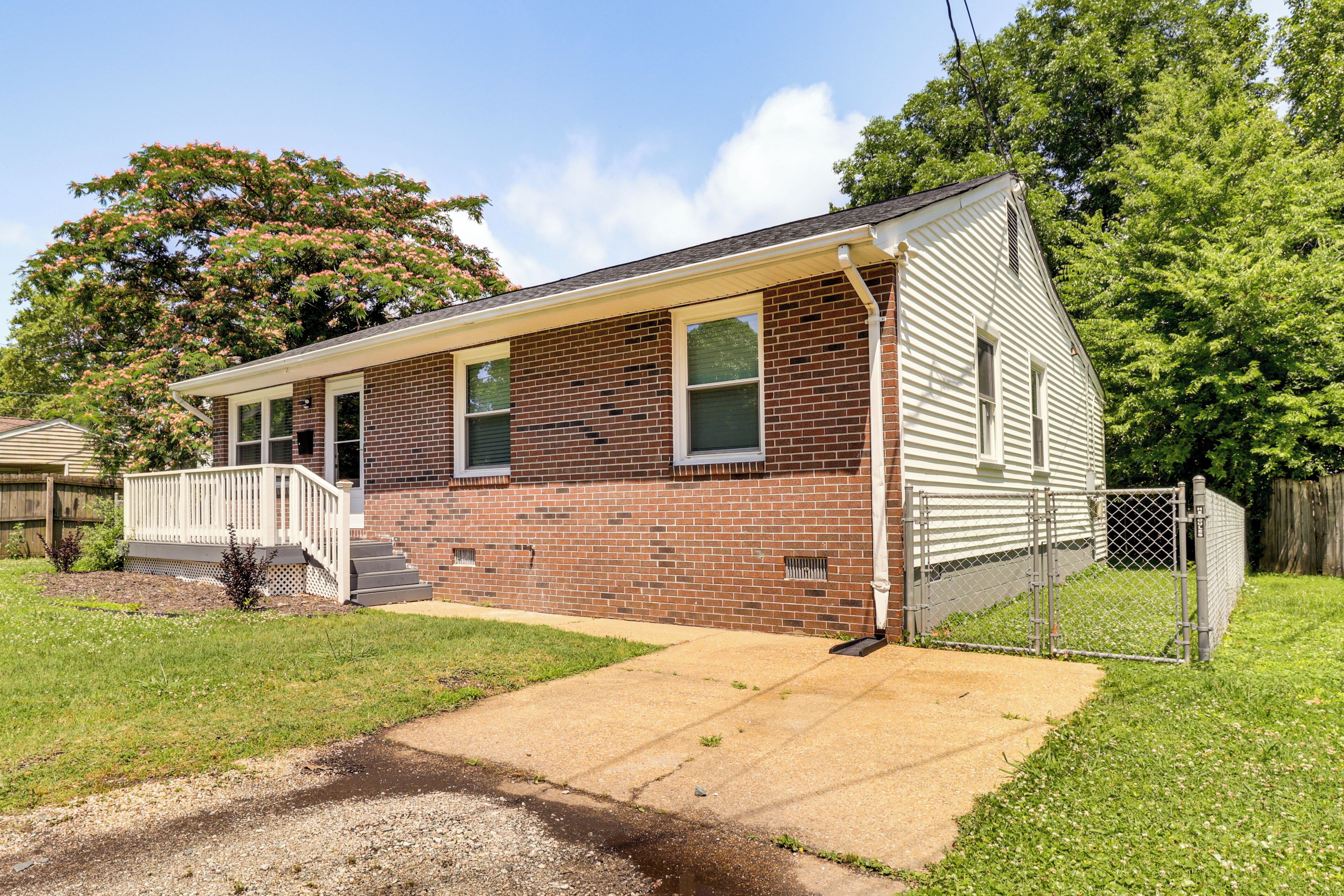A single-story brick house with a front porch, a small front yard, and a chain-link fence.