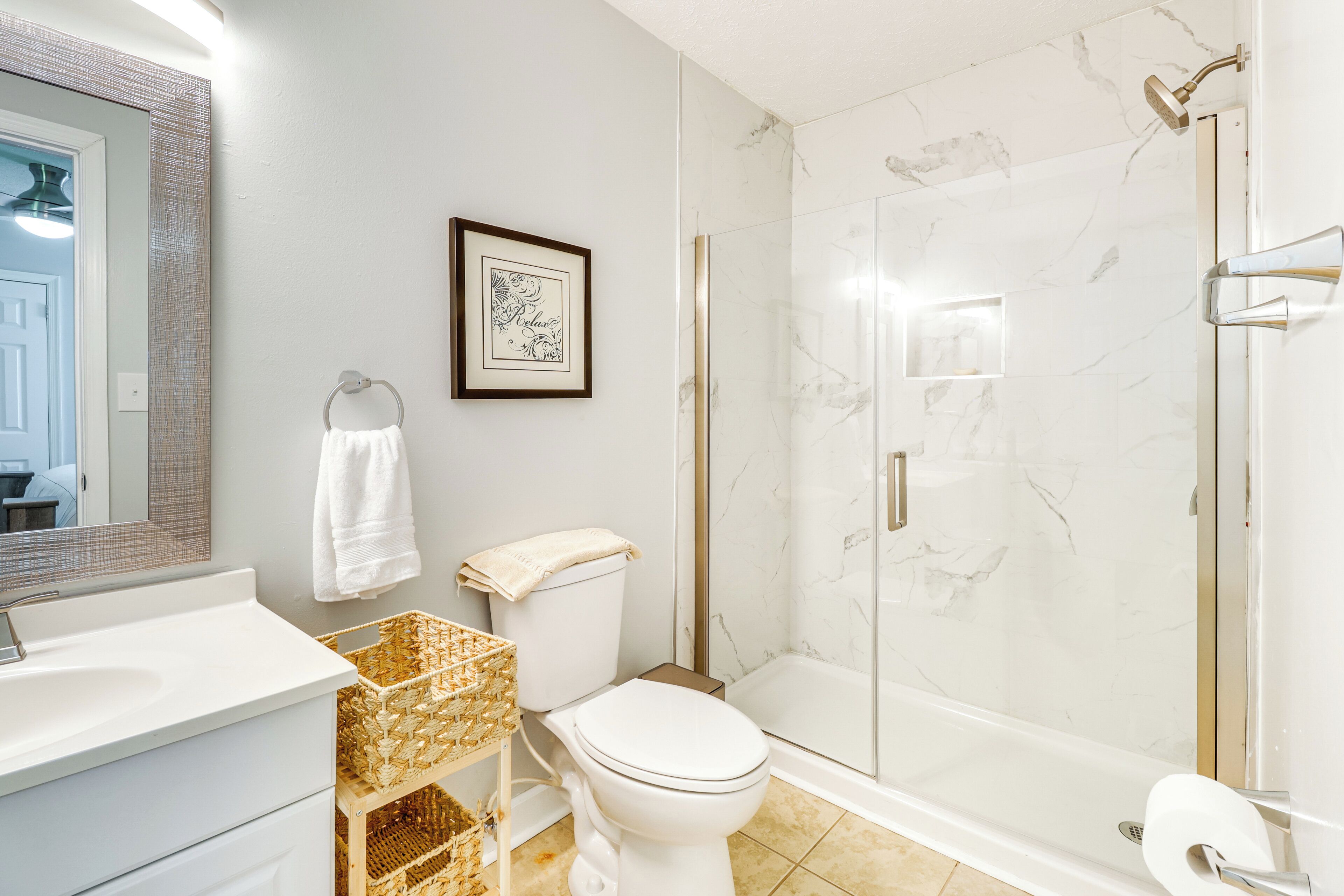 Un bagno con lavandino, wc e doccia bianchi. Si vede un quadro appeso al muro e un cesto sul pavimento.