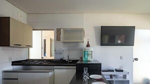 A modern kitchen with a gas stove, white cabinets, a water dispenser, and a flat-screen TV mounted on the wall. - Apartamento em Porto Seguro na Praia de Taperapuã (Porto Seguro)