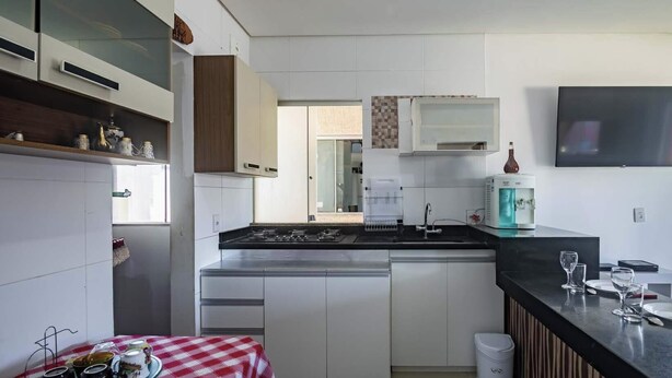 A modern kitchen with white cabinets, a black countertop, and a built-in stove. There is a water dispenser and a flat-screen TV mounted on the wall. - Apartamento em Porto Seguro na Praia de Taperapuã (Porto Seguro)